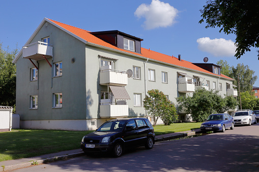 Utsidan av ett ljusgrönt lägenhetshus med vita balkonger och tegelfärgat tak. Framför huset är en liten gräsmatta och sedan en gata där det står bilar parkerade.