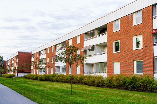 Fasaden av ett lägenhetshus. Fasaden är byggd i rött tegel och det finns vita balkonger. Framför huset finns en häck och sedan gräsmatta.